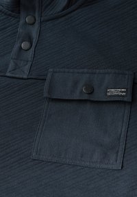 Maglione blu navy con finitura testurizzata, dotato di collo con bottoni, tasca applicata e etichetta con marchio sul lembo della tasca.