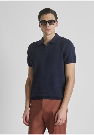 Man met zwarte zonnebril, marineblauw gestructureerd poloshirt met korte mouwen en roestkleurige broek, staand tegen een effen lichte achtergrond.