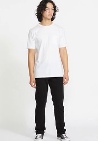 Biele tričko s krátkym rukávom a náprsným vreckom, skombinované s čiernymi nohavicami a čiernymi teniskami, stojace pred jednoduchým bielym pozadím.