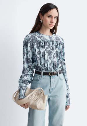 Jonge vrouw draagt lichtblauw paisley blouse, lichtgekleurde hoge taille spijkerbroek met zwarte riem, houdt beige handtas ter hoogte van de taille vast.