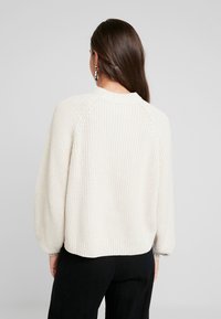 Pull crème en maille épaisse avec un col arrondi, des poignets et un ourlet côtelés, présentant une coupe décontractée et un tissu texturé.