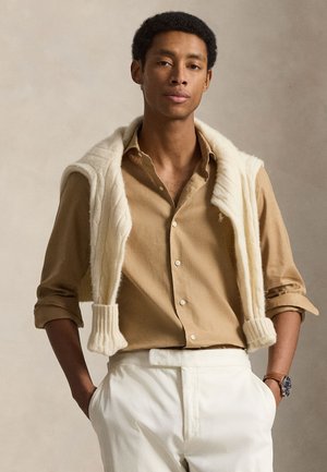 Jeune homme portant une chemise beige à boutons avec les manches retroussées, un pull crème drapé sur les épaules, un pantalon blanc et une montre-bracelet.