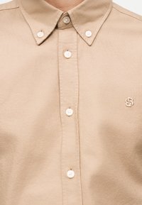 Μπεζ πουκάμισο με κουμπιά λευκούς και ένα μικρό κεντημένο λογότυπο στο αριστερό στήθος, εμφανίζοντας το γιακά και το πάνω μέρος του κορμού.