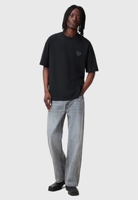 Camiseta negra de manga corta con emblema de corazón en el pecho, combinada con vaqueros de corte holgado de lavado claro y zapatos negros.