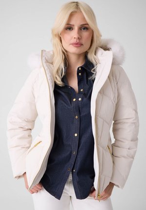 Une femme blonde porte une doudoune blanche bordée de fourrure sur une chemise en jean foncée et un pantalon blanc, devant un fond uni.