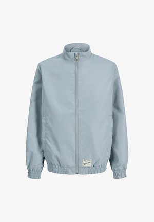 Jongen staande, dragend een lichtblauwe jas met rits, een roze graphic t-shirt, crèmekleurige korte broek, witte sokken en beige schoenen tegen een effen achtergrond.