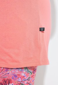 Brzeg koralowo-różowej koszuli z małą czarną metką marki nad kolorowymi spodniami w kwiatowy wzór w odcieniach różu, fioletu i zieleni.