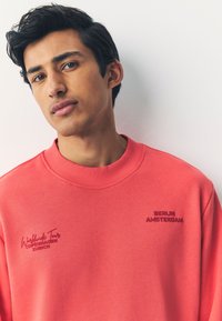 Felpa corallo con collo a girocostina, con testo ricamato: "Worldwide Tour" in rosa scuro e "BERLIN AMSTERDAM" in rosso vivace.