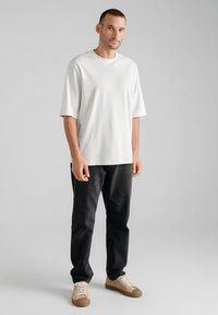 Biała bawełniana koszulka z krótkim rękawem, zestawiona z ciemnymi jeansami denim. Model nosi beżowe trampki z teksturowanymi cholewkami. Prosty i nowoczesny design.