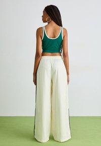 Top verde cropped com acabamentos brancos combinado com calças largas em creme com riscas pretas nas laterais, posicionado contra um fundo claro e um chão verde.