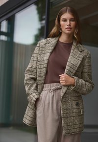 Blazer z texturované hnědé kostkované vlny, s klopovým límcem, přední kapsou a stylovým střihem, spárovaný s hnědým tričkem a šedými kalhotami.