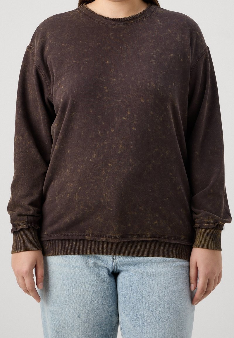 Personne portant un sweat-shirt à manches longues marron foncé délavé et un jean bleu clair, debout devant un fond uni.