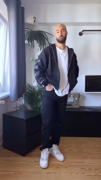 Casaco bomber preto com fecho, t-shirt branca, calças pretas e ténis brancos. O quarto tem piso de madeira e uma planta ao fundo.