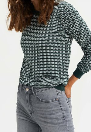 Kvinde iført mørkegrøn og hvid mønstret langærmet top stoppet ned i lysegrå højttaljede jeans med den ene hånd i lommen.
