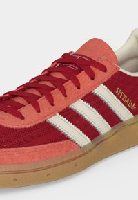 Κόκκινο sneaker Adidas Spezial με λευκές ρίγες, λαστιχένια σόλα και υφή από σουέτ και δίχτυ στο επάνω μέρος, δεμένο με κόκκινα κορδόνια.