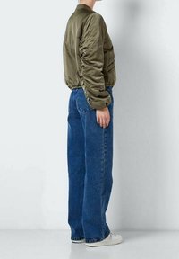 Olivgrön bomberjacka med elastisk nederkant och rynkade ärmar, i kombination med lösa blå jeans och vita sneakers, visad från sidan.