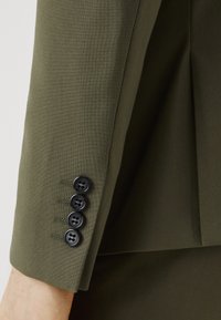 Olive green blazer sleeve featuring a smooth fabric texture and four black buttons along the cuff with contrasting stitching.