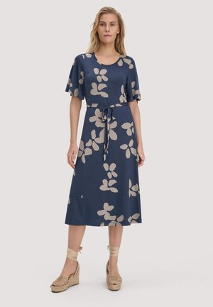 Frau trägt ein marineblaues Midikleid mit beigem abstraktem Blumenmuster, kurzen Ärmeln, Taillengürtel und beigen Keilsandalen mit Knöchelriemen.