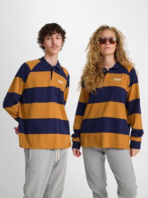 Jeune homme et femme portant des polos à manches longues rayés marron et bleu marine assortis et un pantalon de survêtement gris, debout devant un fond blanc uni.
