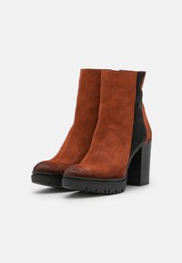 Bottes cheville en daim marron avec un panneau latéral noir, un bout arrondi et un talon noir épais. La semelle est dotée d'une texture rainurée pour une meilleure adhérence.