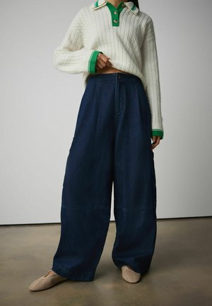 Person wearing a white cable-knit sweater with green collar and cuffs, wide-leg dark blue denim pants, and beige woven slip-on shoes.