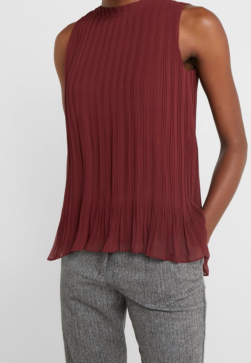 Burgundy plisserad ärmlös topp med hög hals; mjukt, texturerat tyg och fladdrig nederkant. Matchad med grå hundtandsmönstrade byxor.