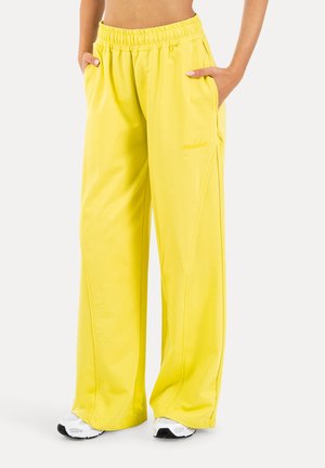 Mujer con pantalones anchos de pierna en color amarillo brillante, con cintura elástica y bolsillos laterales, combinados con zapatillas blancas, manos en los bolsillos.