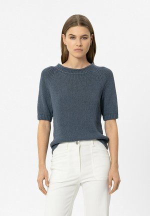 Woman with straight brown hair wearing a blue short-sleeve knit sweater and high-waisted white pants, standing against a plain background.