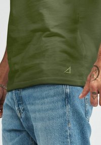 Olivgrön t-shirt med en mjuk textur, med en triangel-logotyp i en ljusare nyans, tillsammans med ljusblå denimjeans.