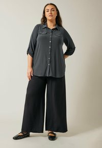 Camisa gris de manga larga con botones en la parte delantera y dos bolsillos en el pecho, combinada con pantalones de pierna ancha negros y zapatos negros con tachuelas.