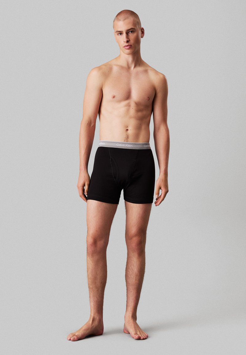 Juodi medvilniniai šortai su pilka Calvin Klein juostele, pasižymintys standartiniu kirpimu ir lygiu paviršiumi. Modelis stovi basomis.