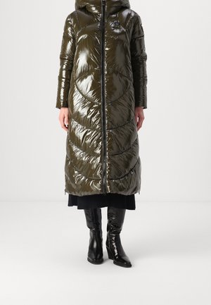 Vrouw draagt een lange, glanzende olijfgroene pufferjas met capuchon, gecombineerd met zwarte laarzen met hak en een zwarte rok.