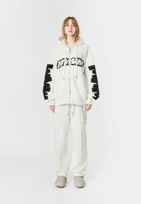 Sudadera blanca de gran tamaño con texto gráfico negro, cierre de cremallera y mangas anchas, combinada con pantalones blancos holgados a juego y zapatos beige texturizados.