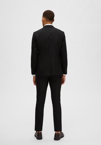 Schwarzer maßgeschneiderter Anzug mit einer Schlitzöffnung, schlank geschnittene Hose und weiße Hemdärmel sichtbar. Braune Schuhe vervollständigen das Ensemble.
