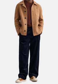 Beige wollen jas met zwarte knopen, een borstzak, gecombineerd met een bruine trui en marineblauwe corduroy broek. Casual sneakers maken de look compleet.