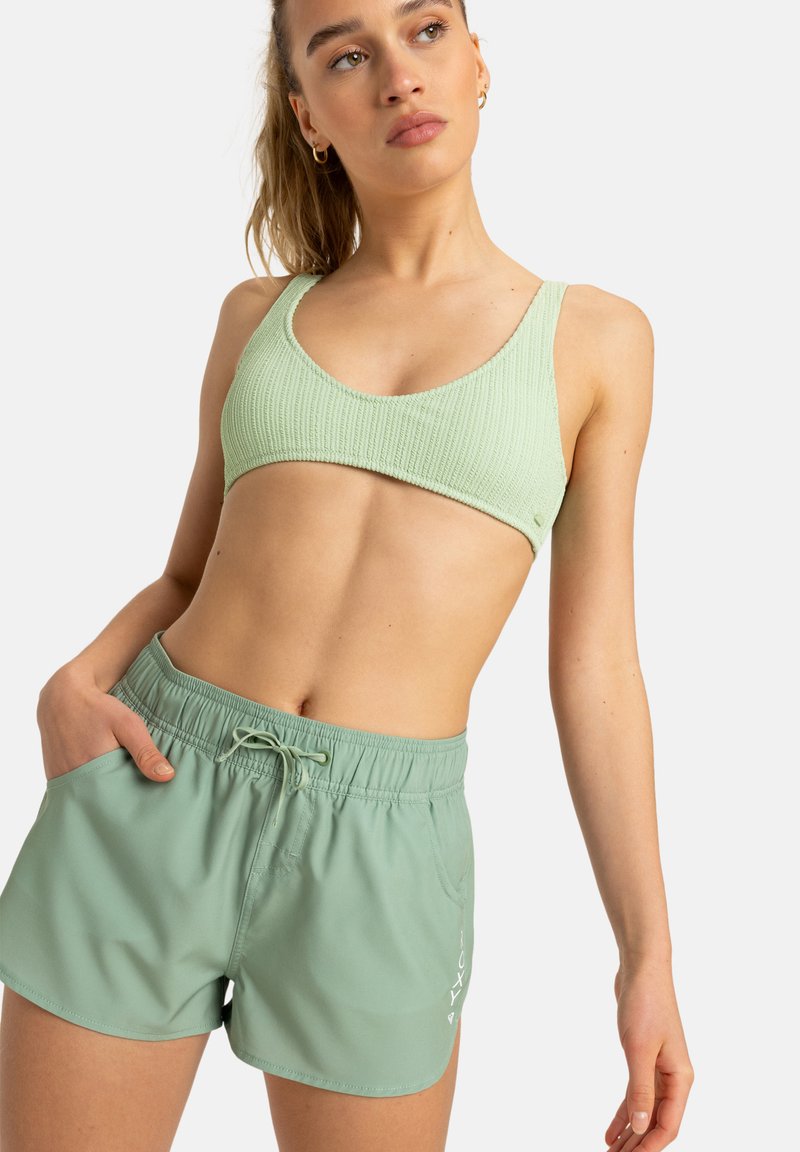 Világoszöld ribbed sportmelltartó széles pántokkal és azonos színű, könnyű shortokkal, amelyek elasztikus derékkal és oldalsó zsebekkel rendelkeznek.