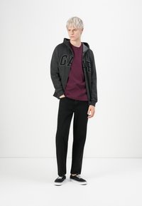 Jeune homme aux cheveux platine portant un hoodie zippé gris foncé de GAP, un t-shirt bordeaux, un pantalon noir et des sneakers noires, se tenant avec les mains dans les poches.