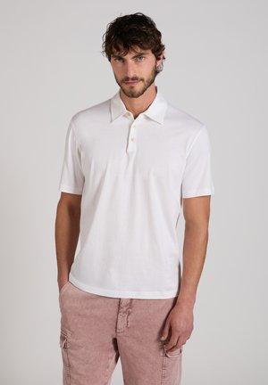 Uomo con i capelli ricci castani che indossa una polo bianca a maniche corte e pantaloni cargo rosa chiaro, in piedi contro uno sfondo semplice.