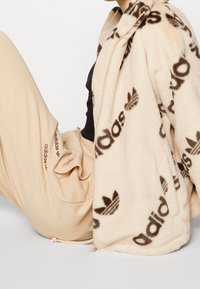 Persona che indossa pantaloni sportivi beige Adidas e una giacca crema soffice con grandi loghi marroni Adidas su uno sfondo bianco semplice.