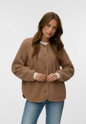 Femme aux longs cheveux bruns portant une veste marron duveteuse boutonnée et un jean bleu, debout devant un fond clair uni.