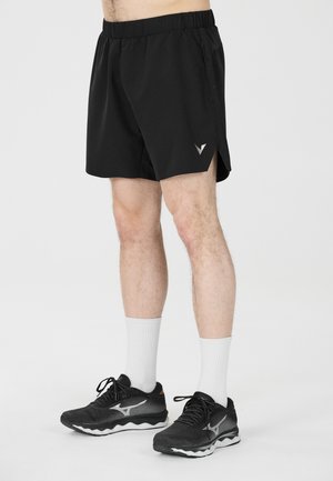 Mannelijke benen met zwarte sportshorts met een klein logo, witte sportsokken en zwarte hardloopschoenen op een lichte achtergrond.
