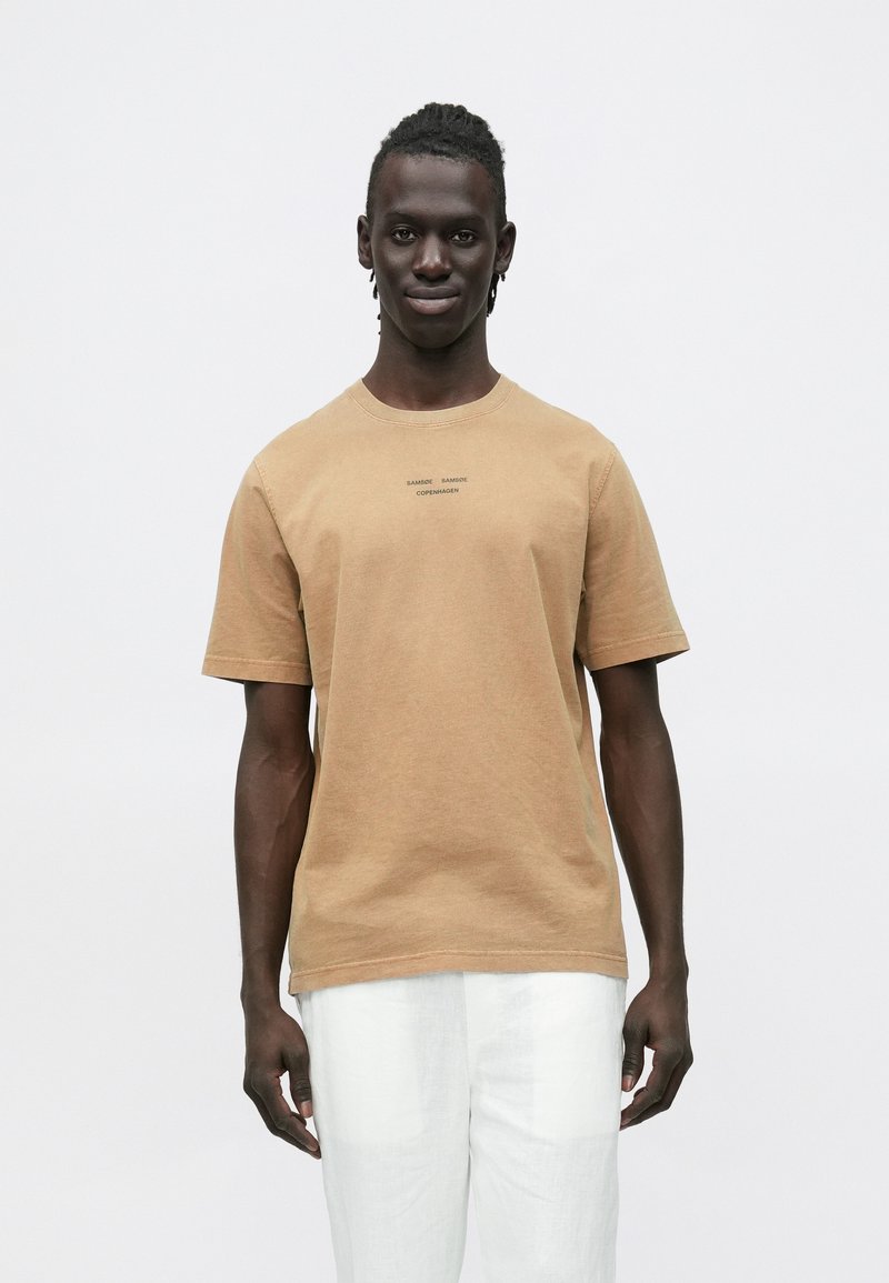 T-shirt de algodão castanha com mangas curtas, gola redonda e texto impresso subtil no peito. Textura suave com um corte descontraído.