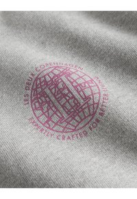 Szary materiał o teksturowanej powierzchni z okrągłym fioletowym logo. Logo zawiera napis "Les Deux Copenhagen" oraz wzór w kratkę.