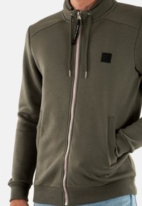 Felpa con zip verde oliva, collo alto, tasche laterali, polsini a costine e zip a contrasto. Patch con logo nero sul petto.