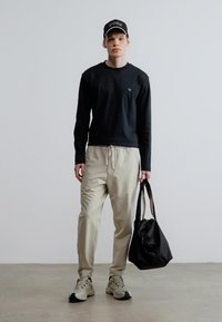 Fekete hosszú ujjú póló, bézs színű húzózsinóros nadrág, bézs sportcipők és egy fekete táska. A sapkán logó látható, a modell semleges háttér előtt áll.