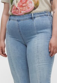 Person iført lyseblå højttaljede jeans og en beige skjorte med en stor pink grapefrugtgrafik, stående med afslappede arme.