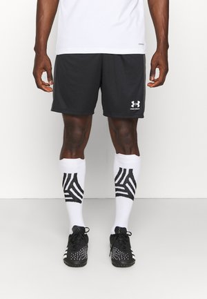 Pantalones cortos deportivos negros con el logo blanco de Under Armour, combinados con calcetas blancas que presentan un patrón geométrico negro y zapatos deportivos negros con acentos blancos.