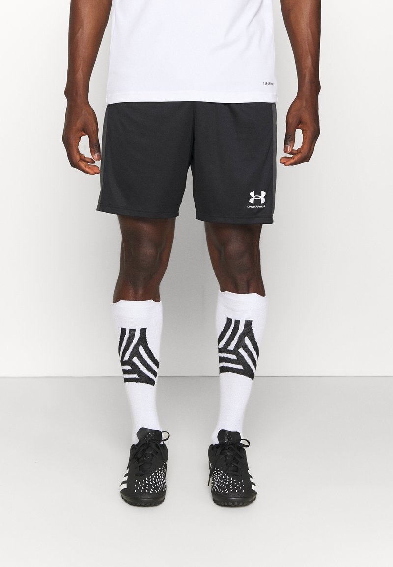 Svarta träningsshorts med vitt Under Armour-logotyp, i kombination med vita strumpor med svart geometrisk mönster och svarta sportskor med vita detaljer.