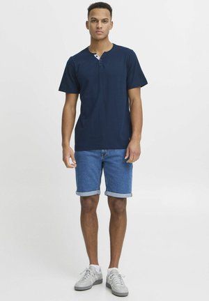 Camisa henley de manga corta en azul marino con tapeta de tres botones, combinada con pantalones cortos de mezclilla con dobladillo enrollado y zapatillas grises. Diseño simple, ajuste casual.