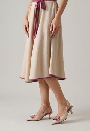 Kobieta w beżowej spódnicy midi z fioletowym obszyciem i beżowych szpilkach typu slingback z kolorową kokardą, stojąca na jednolitym tle.